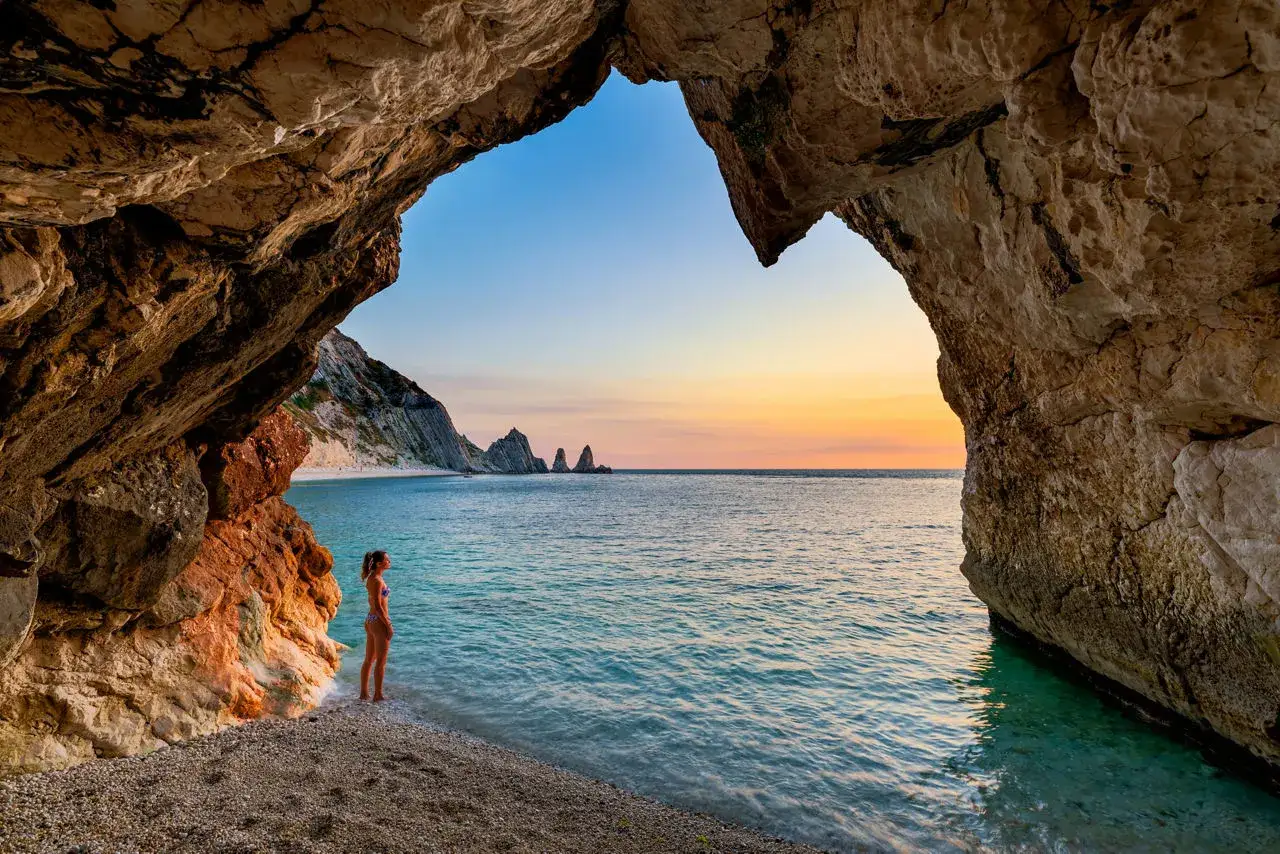 Una ragazza ammira il tramonto da una grotta sulle spiagge del Conero. Il mare turchese lambisce la riva di ciottoli.