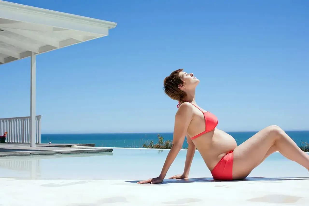 Donna incinta in bikini rosso si gode il sole vicino a una piscina a sfioro con il mare sullo sfondo. Un momento di pace per il suo mare in gravidanza.