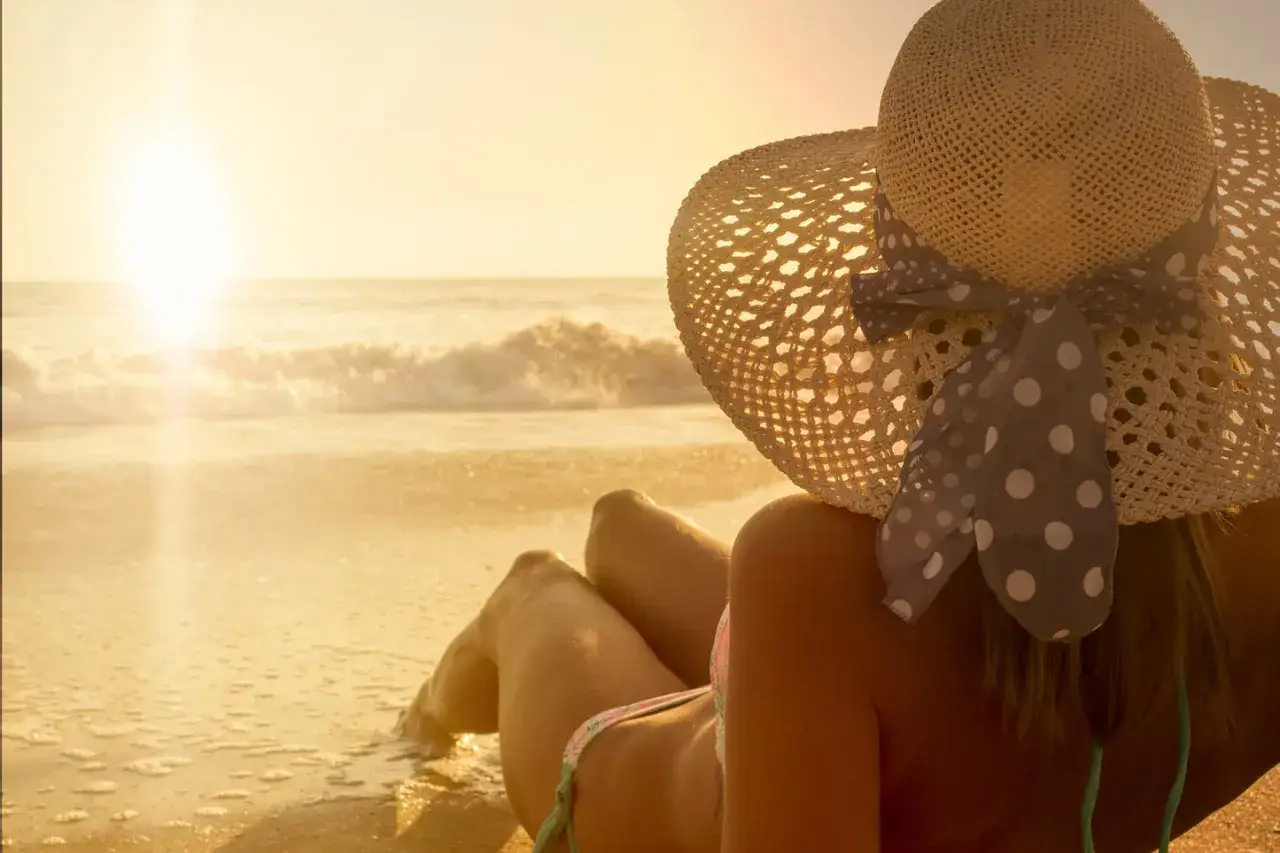 Donna con cappello di paglia e un'abbronzatura scura, sdraiata sulla spiaggia dorata al tramonto.