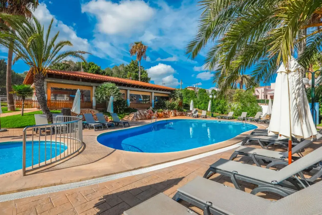 Piscina azzurra e lettini prendisole al hotel Occidental Menorca, con palme e cielo azzurro.
