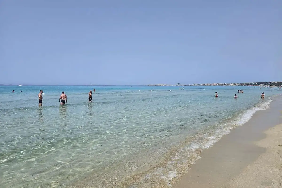 Persone godono del limpido mare di Gallipoli, con la costa in lontananza sotto un cielo azzurro.