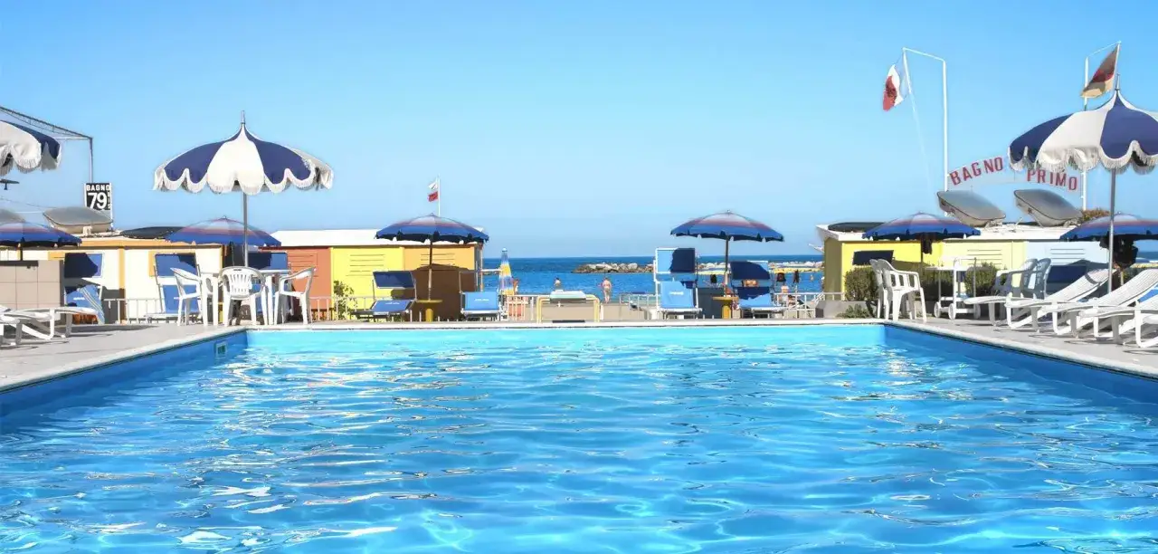 Piscina azzurra e lettini prendisole al Valentini Family Village, con cabine colorate e il mare sullo sfondo.