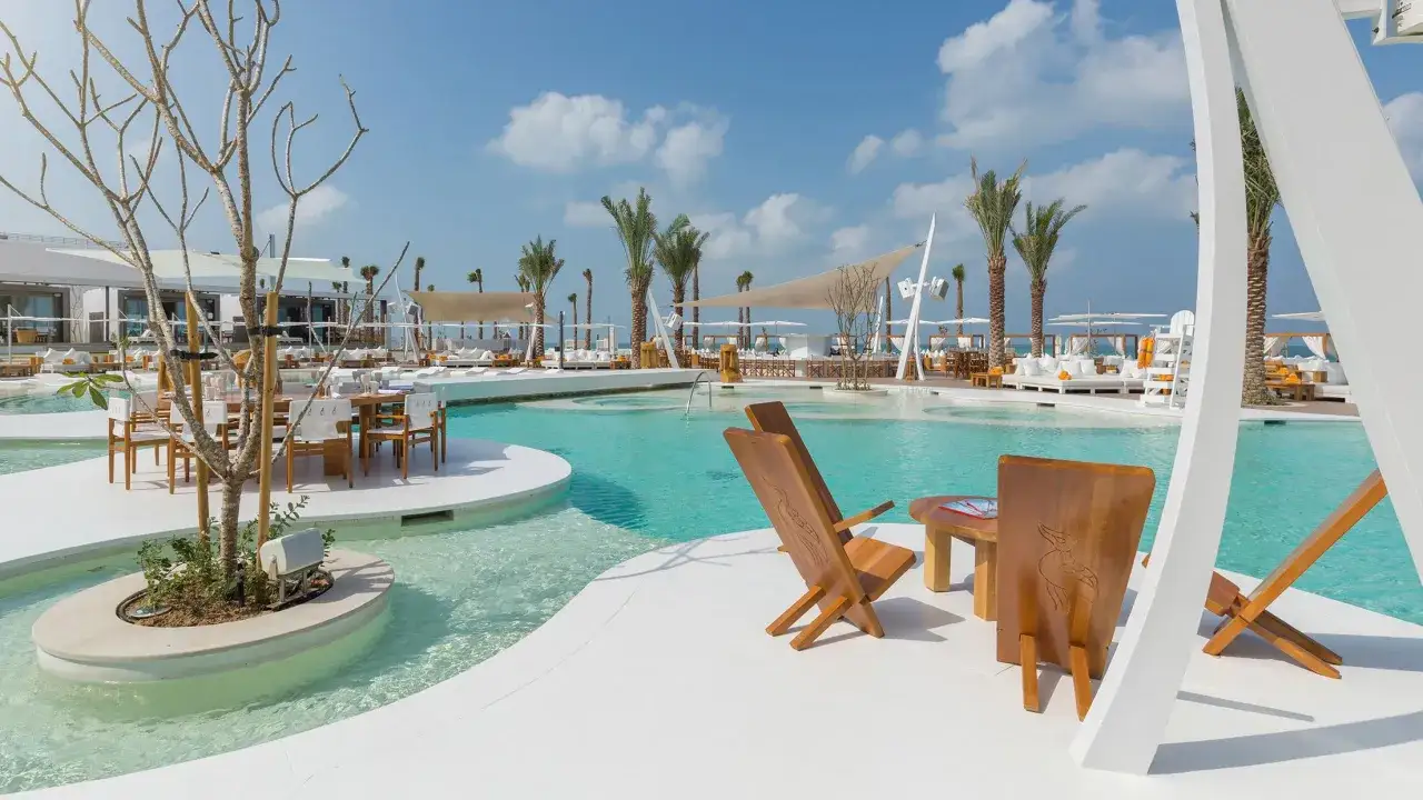 Lussuoso beach club a Dubai con piscina turchese, lettini bianchi, palme e tavoli apparecchiati per un'esperienza indimenticabile.