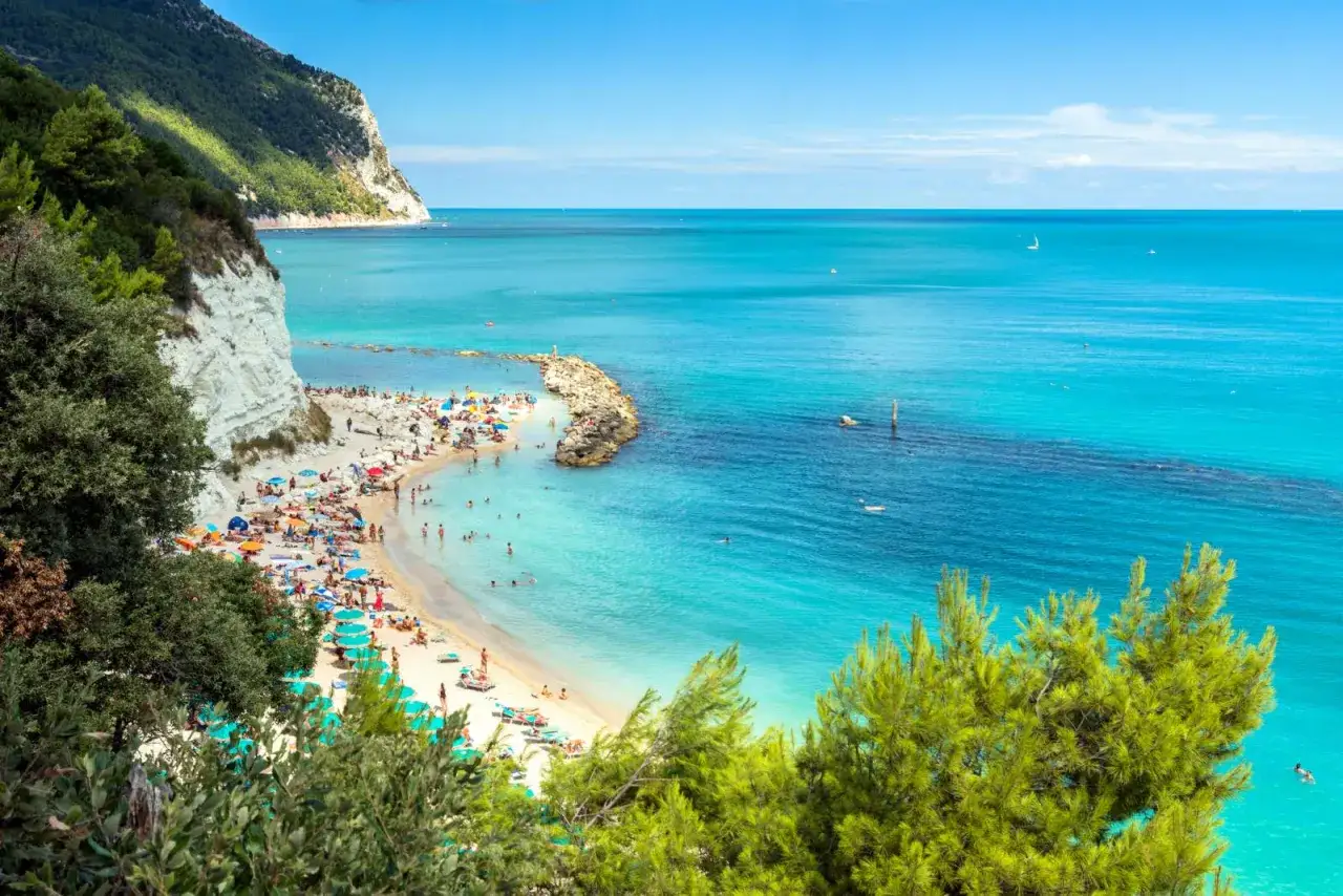 Spiaggia affollata con ombrelloni colorati e gente che nuota nel mare cristallino dell'Abruzzo. Scogliere bianche e vegetazione rigogliosa incorniciano questo paradiso.