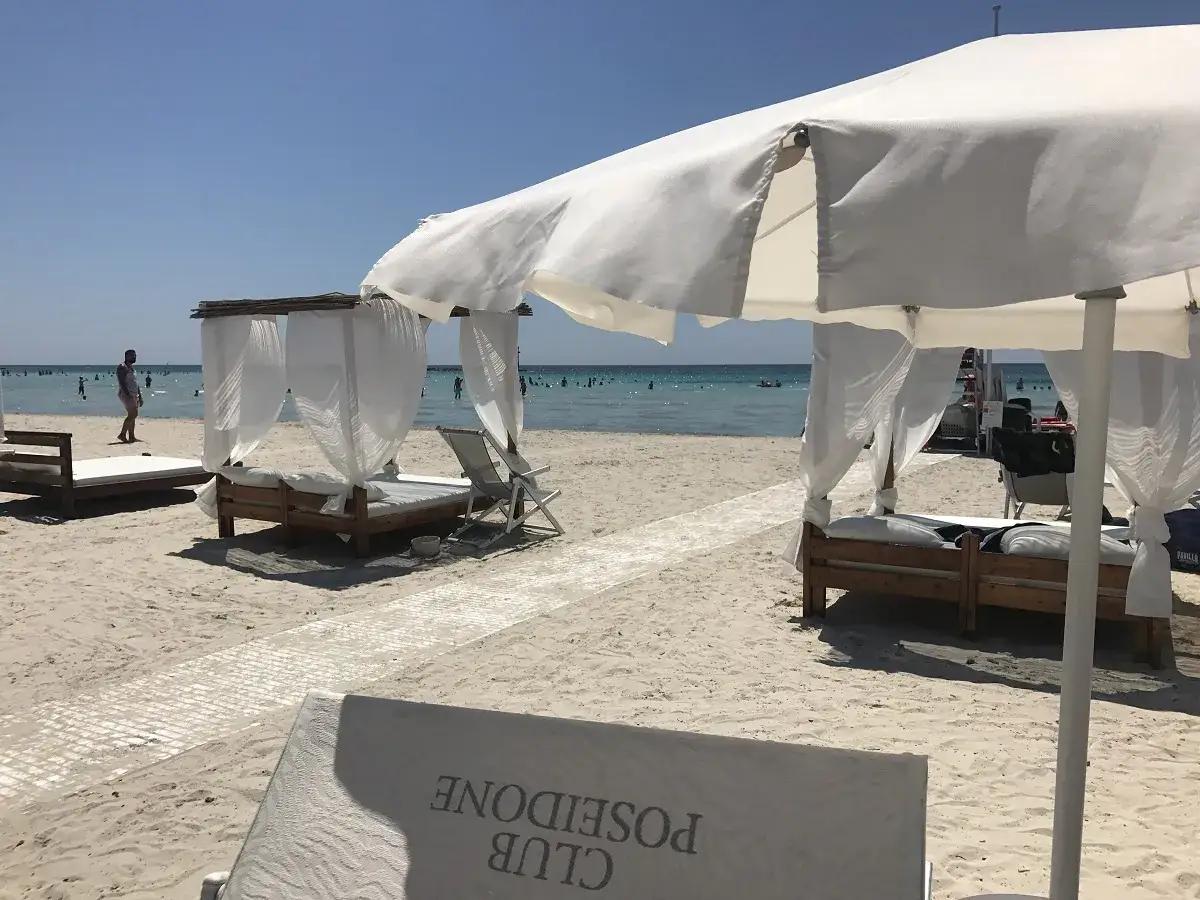 Lettini e gazebo bianchi sulla sabbia del Poseidone Beach Resort Ugento, con il mare azzurro e gente che nuota sullo sfondo.