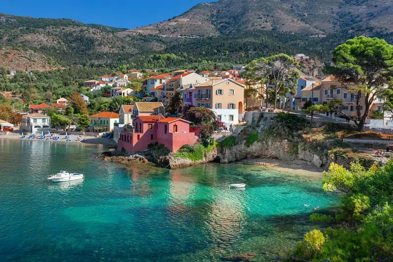 Case colorate affacciate su acque cristalline, un sogno di spiagge Grecia. Barche a vela e colline verdi completano il quadro.